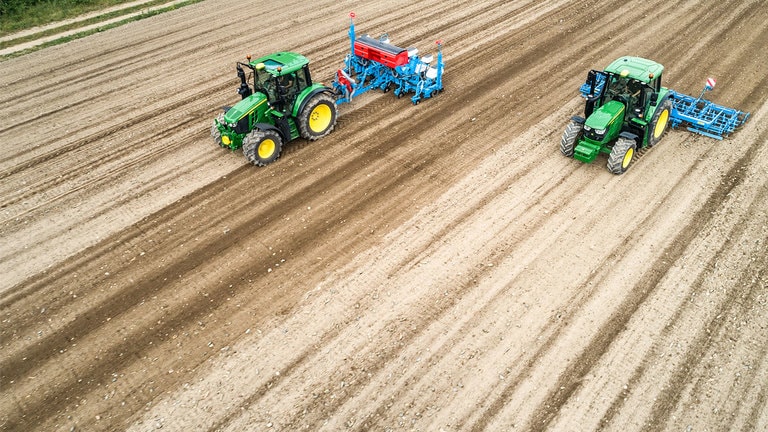 Kaks John Deere'i 6M traktorid paljal põllul. Üks veab Monsomen'i külvikut. Teine veab Lemken'i peenrakombaini