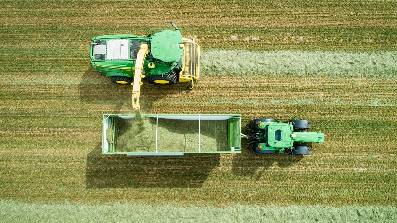 Aerofoto John Deere 8000-seeria liikurhekseldist koos murukoguriga, mis laadib silo John Deere traktori poolt veetavasse kärusse