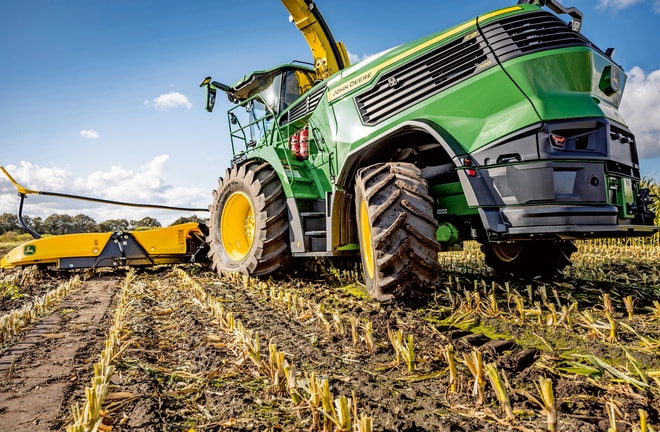 John Deere'i hekseldi töötab koristatud maisipõllul, näidates masina suuri rehve ja lõikemehhanismi selge sinise taeva taustal.